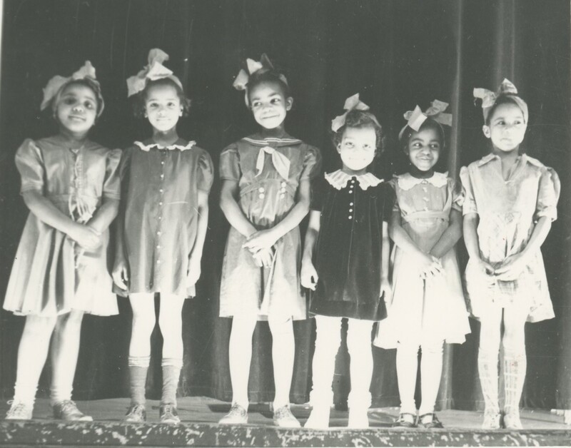 Six little children stood shoulder to should on a stage for a performance all wearing dresses and a bow on their head.