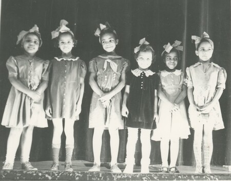 Six little children stood shoulder to should on a stage for a performance all wearing dresses and a bow on their head.